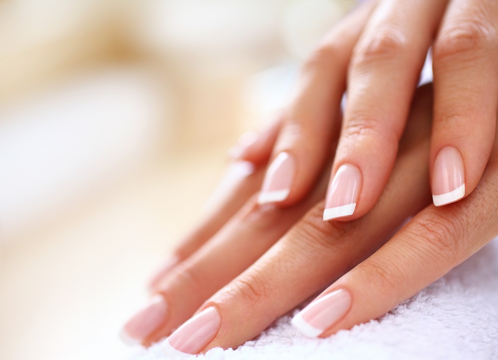 Closeup of nicely manicured female fingernails. One hand is placed on top of other, both on a white towel. Very nice french manicure with transparent nail paint. Blurry beige background. Copy space.