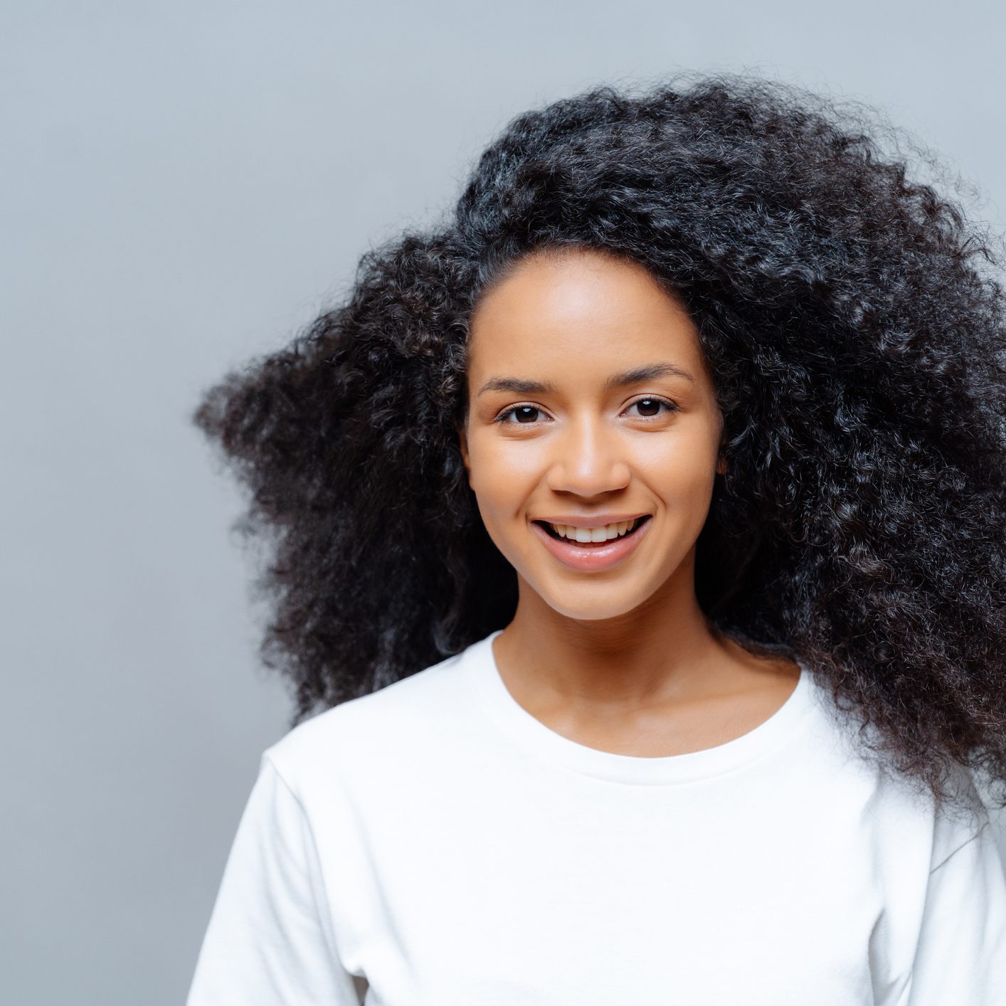 Positive curly woman with natural beauty, dressed in white casual t shirt, has happy expression, looks directly at camera, poses against grey background. Cheerful teenage girl expresses good emotions