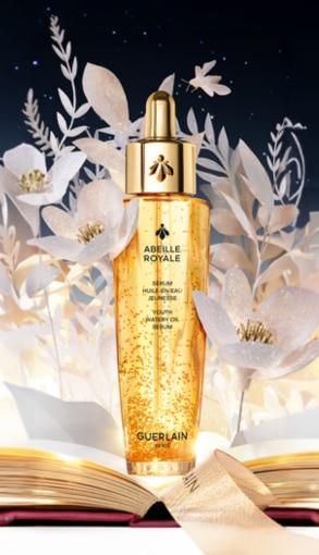 Flacone Guerlain Abeille Royale olio viso dorato con particelle luminose, su sfondo floreale bianco e libro aperto, atmosfera elegante.
