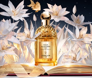 Flacone Guerlain Aqua Allegoria dorato su sfondo floreale bianco con libro aperto e decorazioni eleganti.