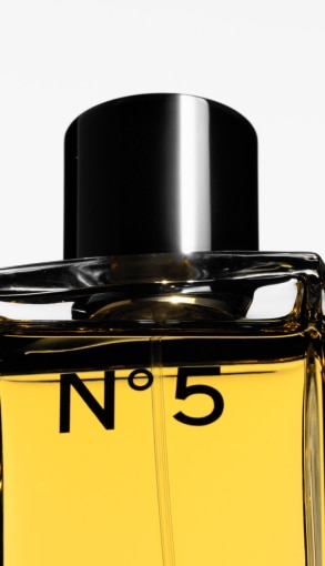 Dettaglio ravvicinato del flacone Chanel N°5 con liquido ambrato e tappo nero lucido su sfondo chiaro.