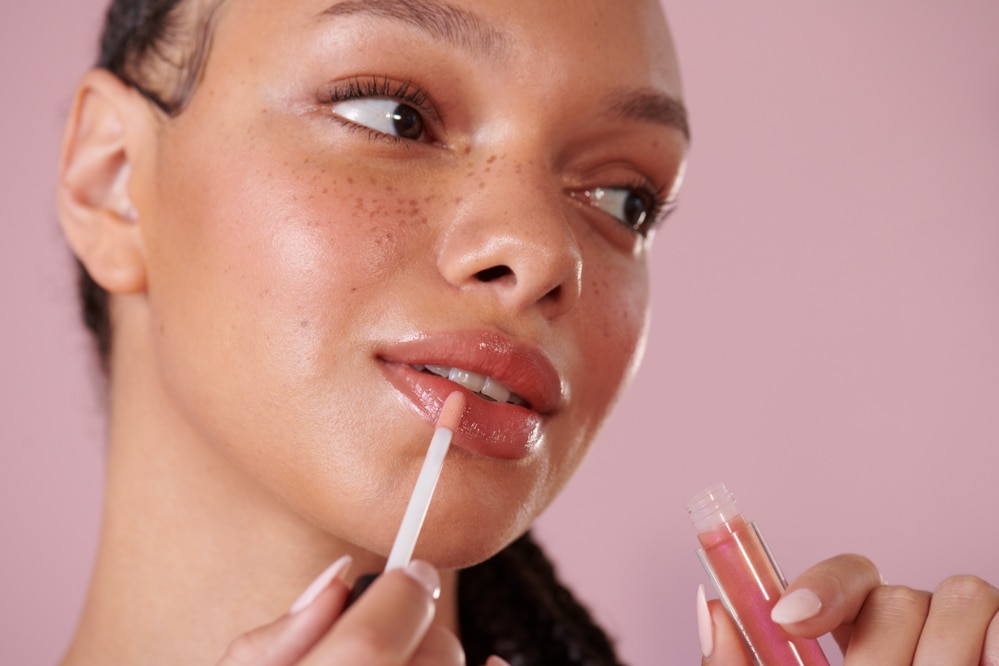 Mano che applica gloss labbra rosa con sfondo rosa tenue.