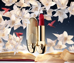 Rossetto Guerlain dorato e rosso su sfondo floreale bianco con libro aperto e decorazioni eleganti.