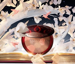 Guerlain Terracotta cipria illuminante con fiori bianchi e nastro dorato su sfondo artistico.
