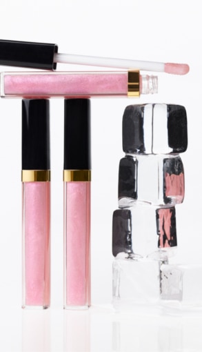 CHANEL gloss labbra rosa chiaro con applicatore, flaconi su superficie riflettente accanto a cubi trasparenti lucidi
